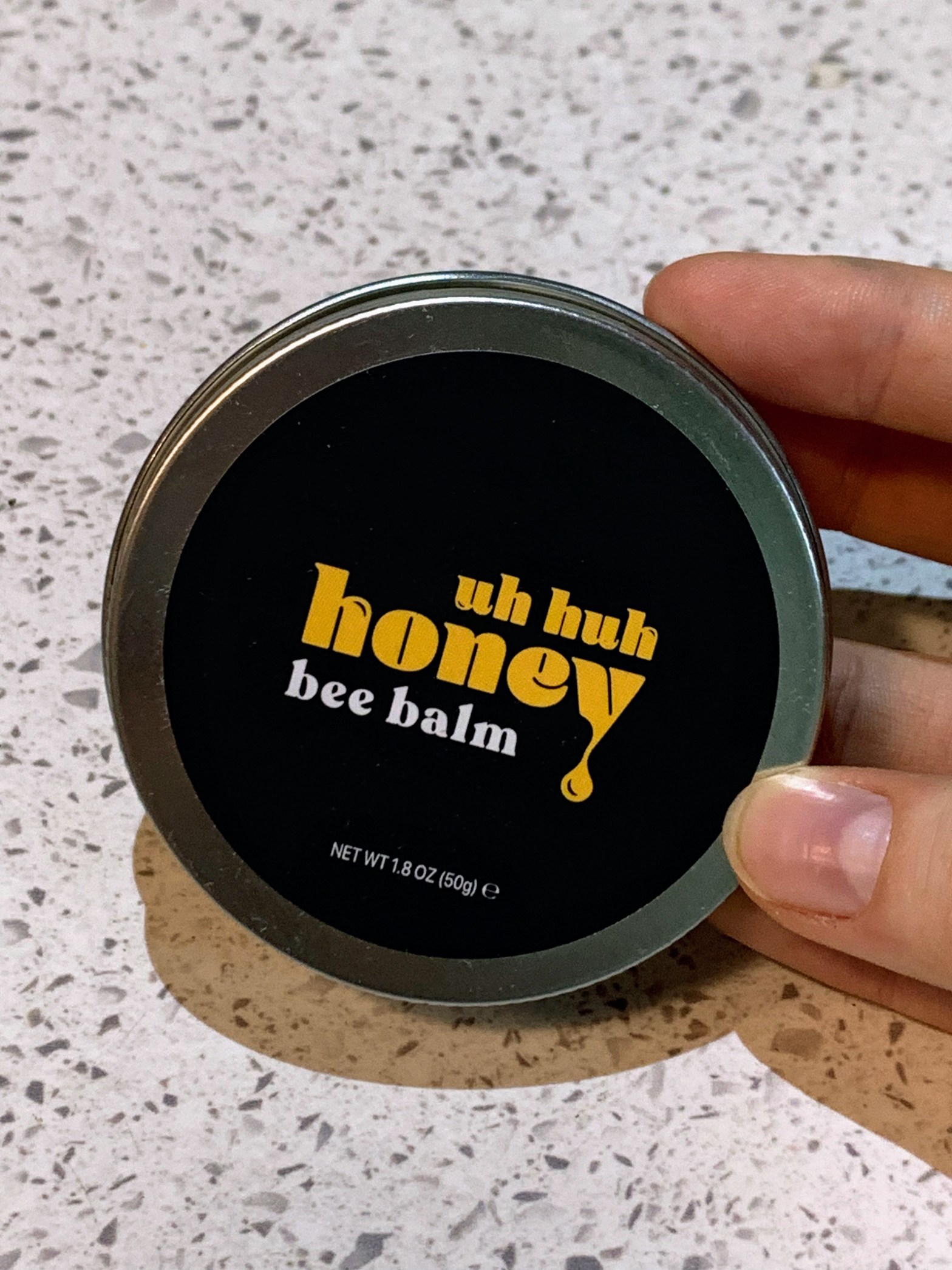 A small metal tin with a black label that reads "Uh Huh Honey Bee Balm". Held in a Caucasian hand against a white countertop